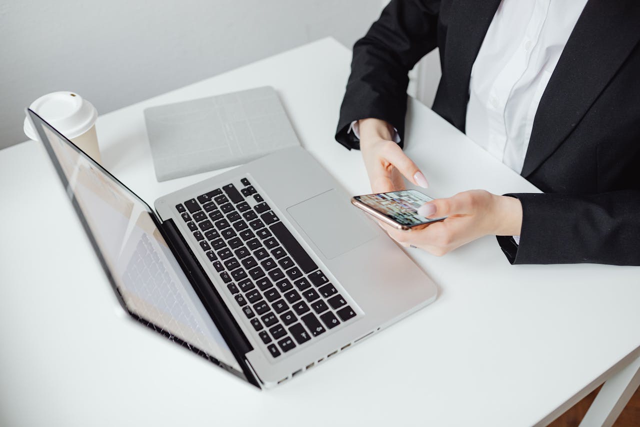 Businesswoman using smartphone at desk with laptop and coffee cup.